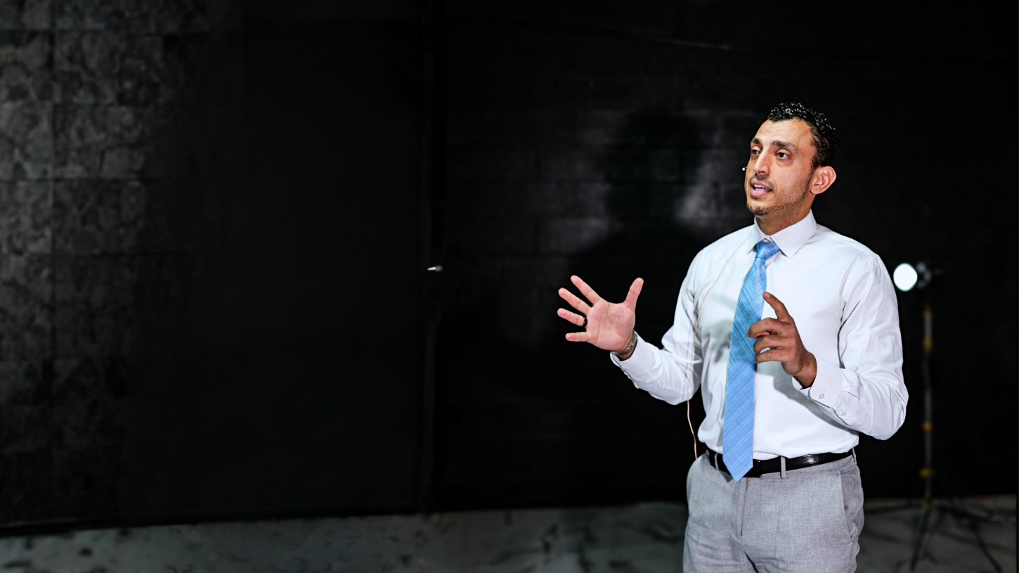 Ibrahim J. Awad speaking on stage, mid-gesture, wearing a lapel mic