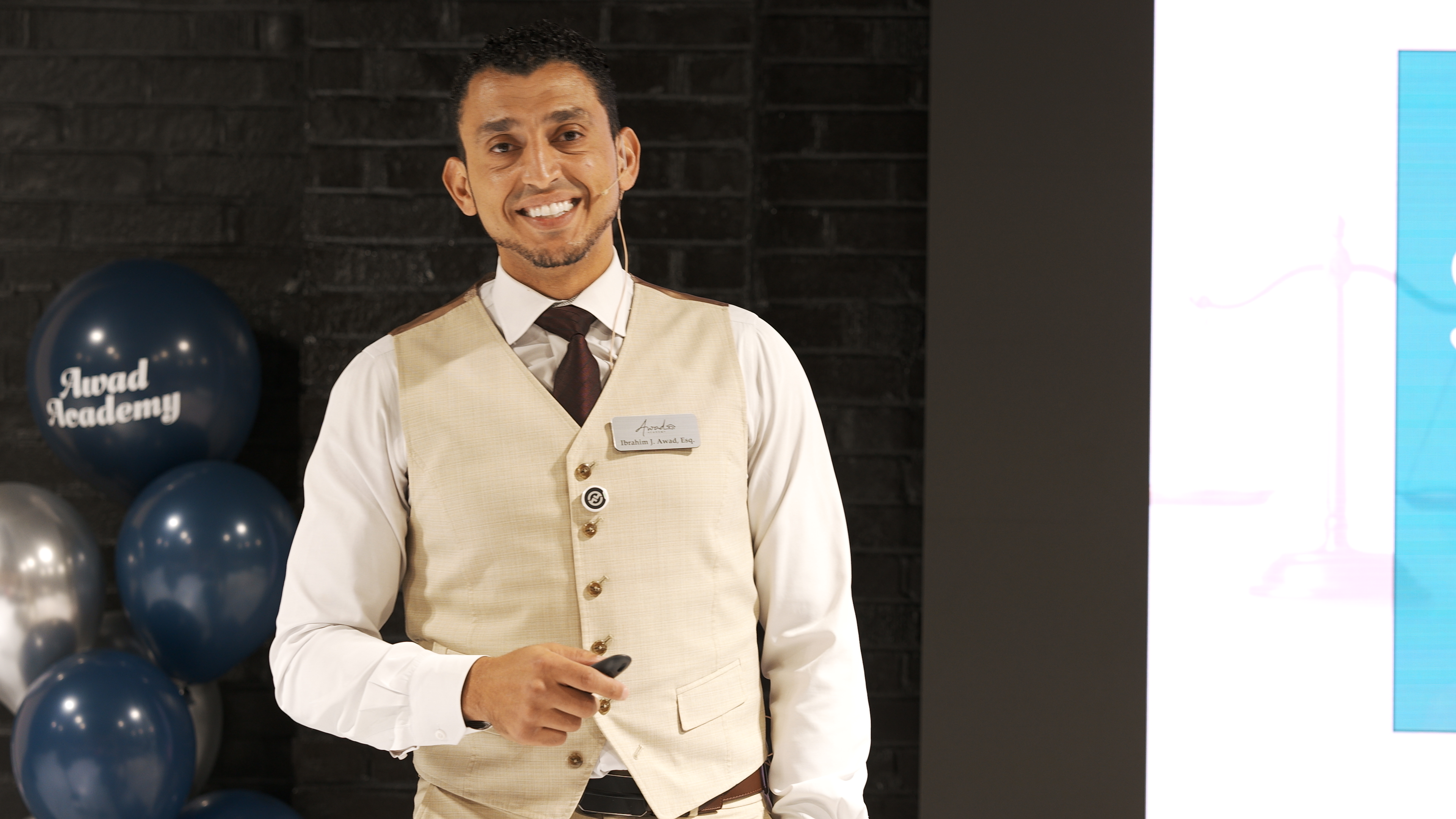Ibrahim J. Awad presenting on stage in a waistcoat and headset microphone, Awad Academy balloons beside him