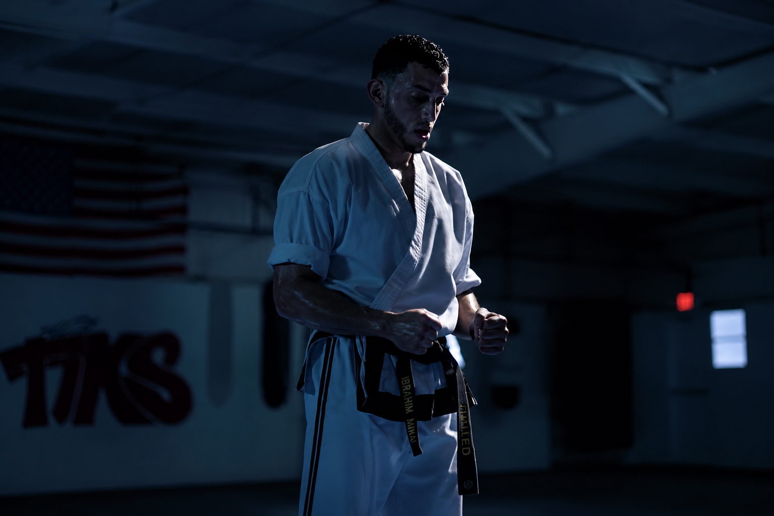 Ibrahim J. Awad in a gi and black belt, hands up, in a dimly lit dojo