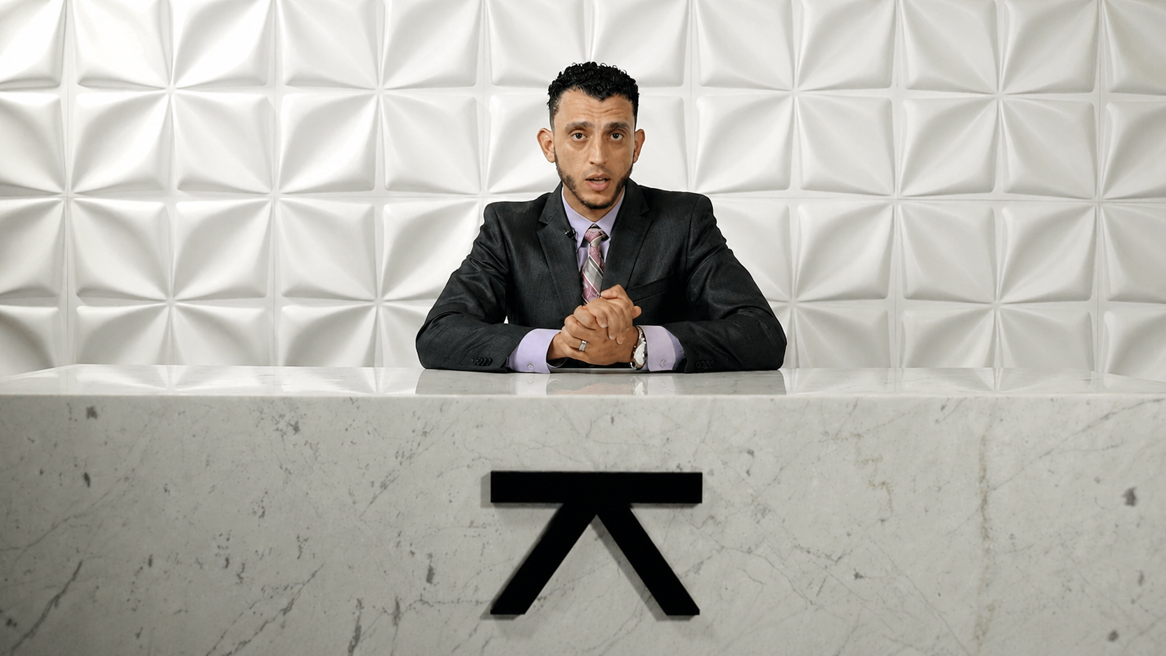 Ibrahim J. Awad seated at a marble desk in front of a white panelled wall, mid-piece to camera